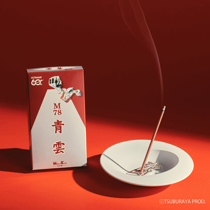 A red and white box of incense stands beside a white dish holding a single lit incense stick with smoke rising against a deep red background.