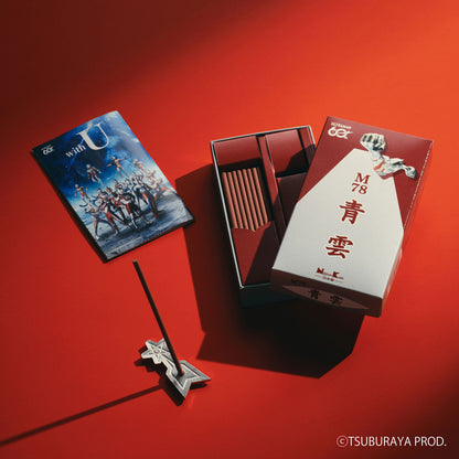 A styled product shot of a matchbox-style incense set on a vivid red background. The set includes a rectangular box with a white and red front, an open inner box showing reddish incense sticks, a printed insert card with illustrated figures, and a silver star-shaped incense holder with a stick resting in it.